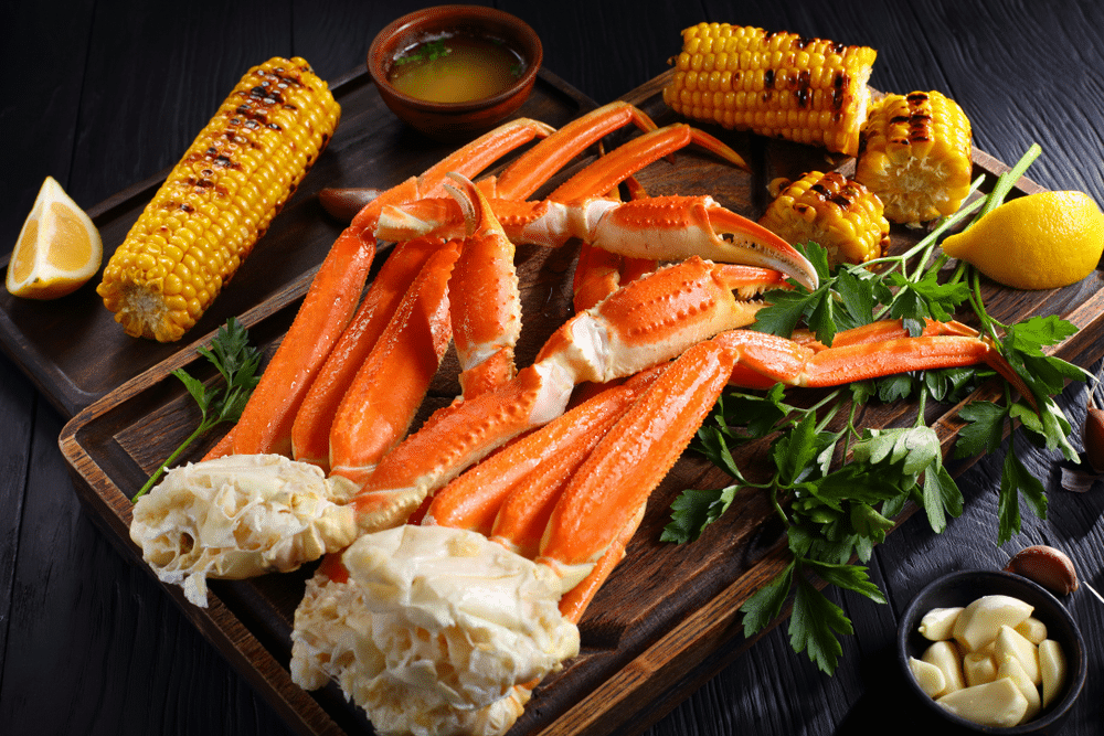 snow Crab legs served with melted butter garlic cloves lemon slices grilled corn in cobs and fresh parsley on wooden cutting board